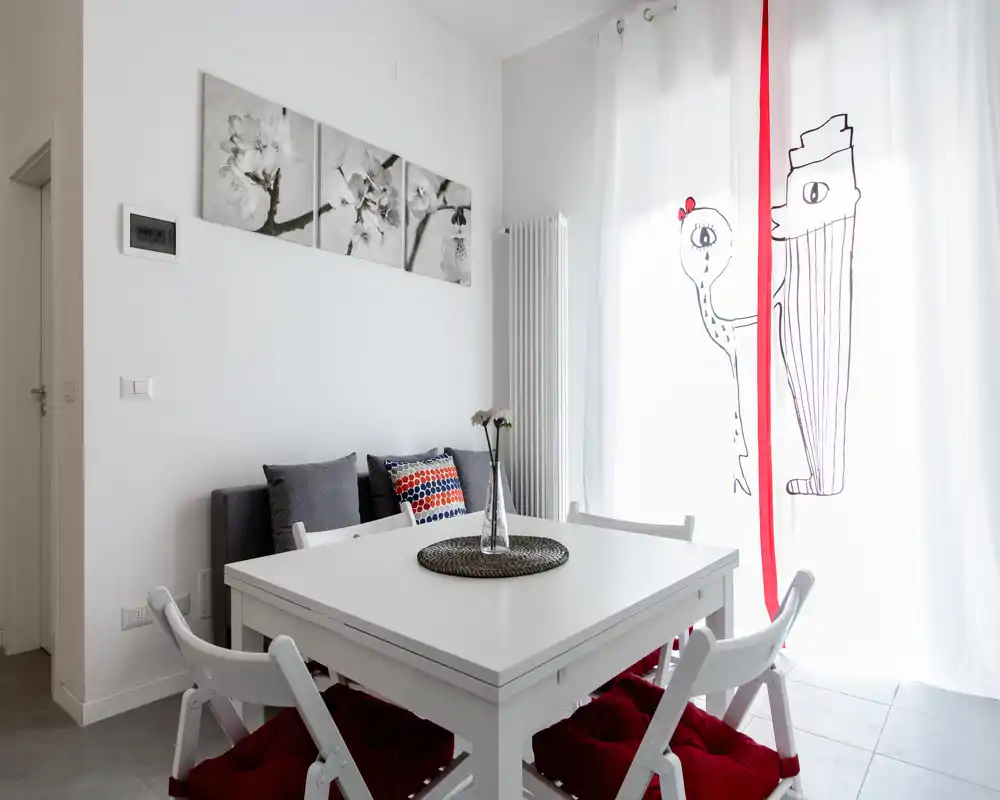 Zona pranzo luminosa e minimal con un tavolo bianco, sedie pieghevoli imbottite e cuscini decorativi accanto a una finestra luminosa con tende leggere.