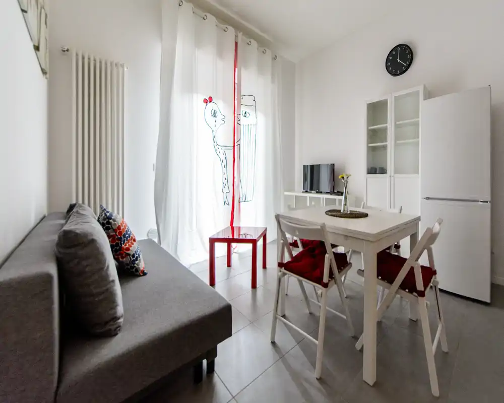 Zona giorno luminosa e compatta con divano grigio, tavolo da pranzo per quattro e una piccola TV — arredamento pulito e tocchi rossi che rendono l'ambiente accogliente.