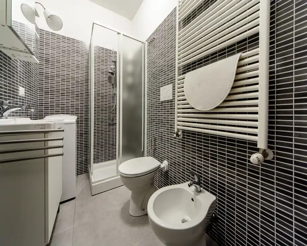 Bagno moderno piastrellato con cabina doccia, WC e bidet, sanitari bianchi e scalda salviette. Ambiente pulito, luminoso e funzionale, adatto per affitti brevi.