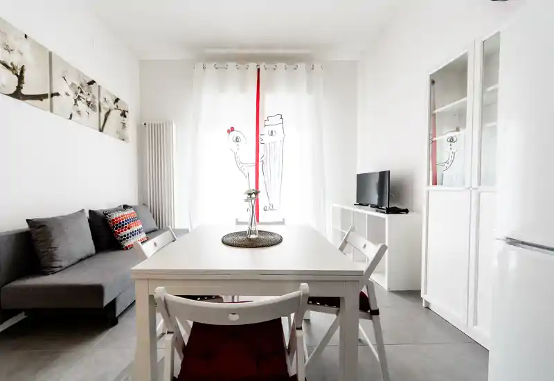 Luminoso soggiorno con area pranzo moderna: tavolo bianco centrale, divano grigio con cuscini decorativi; arredo minimal e molta luce naturale rendono l'ambiente arioso.