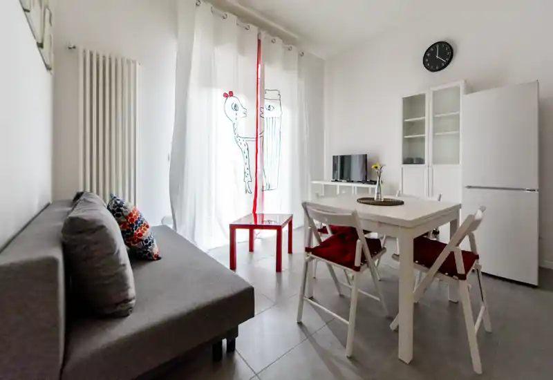 Zona giorno luminosa e compatta con divano grigio, tavolo da pranzo per quattro e una piccola TV — arredamento pulito e tocchi rossi che rendono l'ambiente accogliente.
