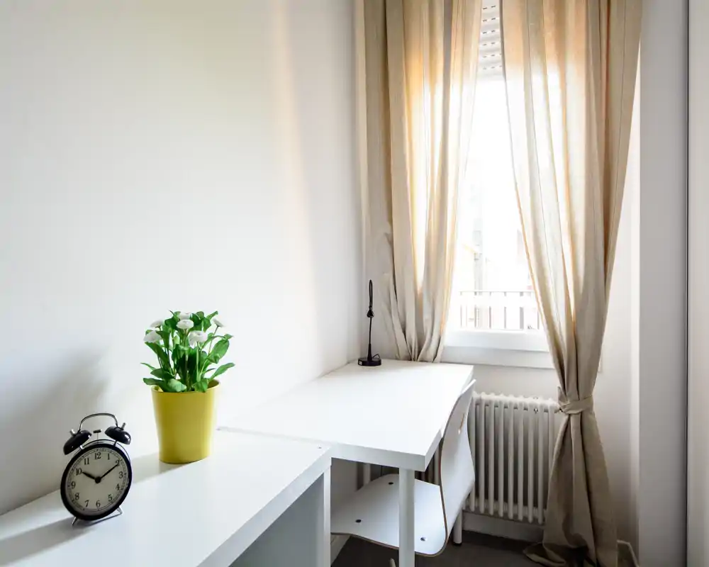 Piccolo spazio di lavoro luminoso vicino alla finestra con scrivania bianca, sedia, lampada e pianta in vaso — minimal, pulito e arioso.