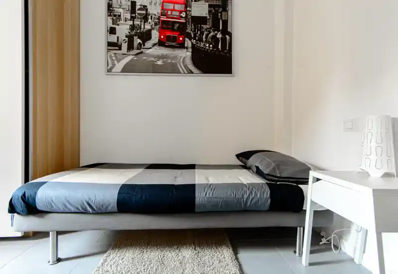 Camera da letto singola moderna e luminosa con letto rifatto, comodino minimale e quadro contemporaneo sopra la testiera. Linee pulite, toni neutri e un piccolo tappeto creano uno spazio riposante e funzionale ideale per soggiorni brevi.