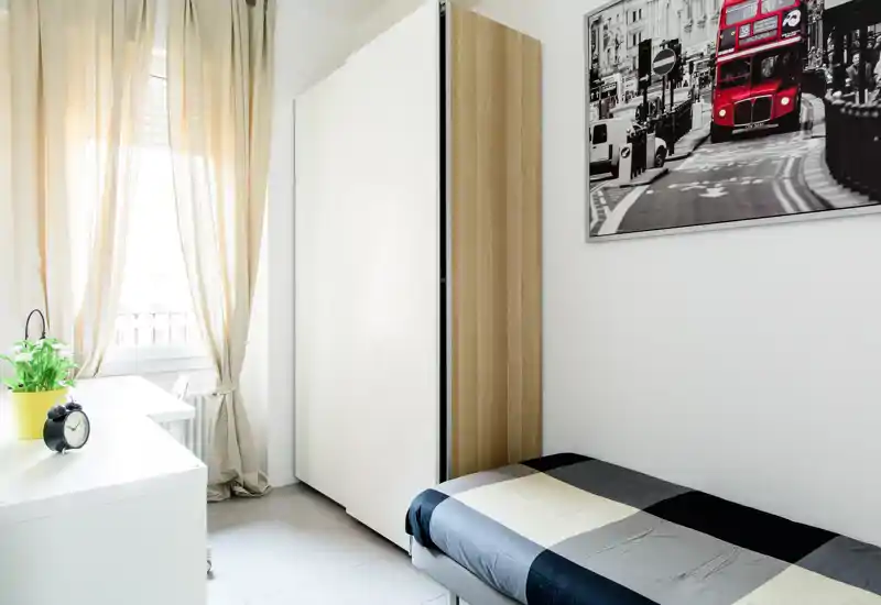 Camera da letto compatta e luminosa con letto singolo, armadio e scrivania vicino alla finestra; arredo moderno e minimal con luce naturale.