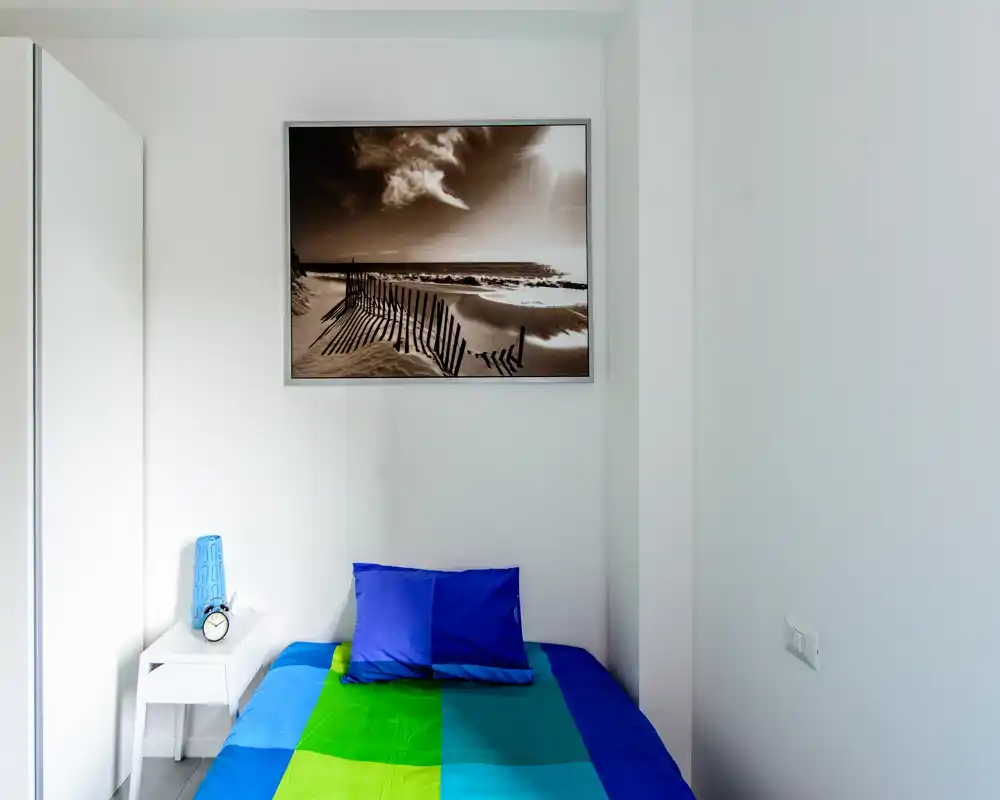 Petite chambre lumineuse avec un lit simple habillé de linge bleu et vert, une table de chevet avec lampe et réveil, et un tableau accroché au-dessus de la tête de lit.