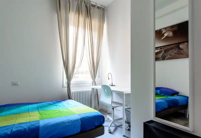 Chambre lumineuse et soignée avec lit aux draps colorés, petit bureau avec lampe près de la fenêtre et un miroir pleine hauteur reflétant une partie du lit.