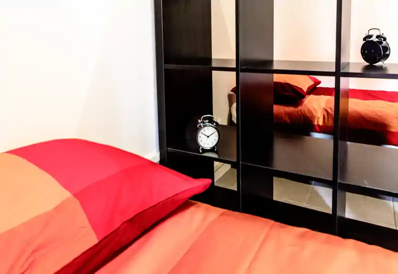 Camera da letto luminosa e accogliente con una coperta rosso-arancione, cuscini e un mobile a scomparti che riflette una sveglia.