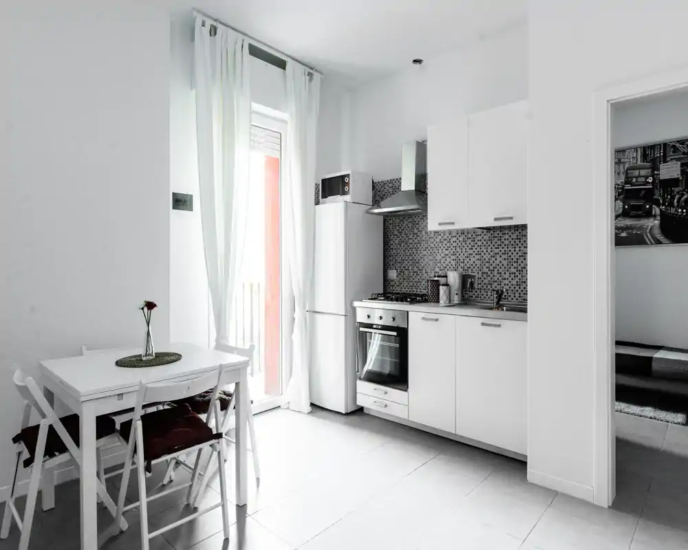 Cucina compatta e luminosa con forno integrato, frigorifero e un piccolo tavolo da pranzo vicino alla porta del balcone — pensili bianchi e paraschizzi a mosaico per un ambiente pulito e accogliente.