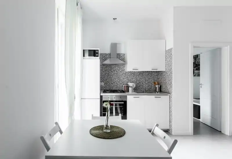 Cucinotto moderno e luminoso con elettrodomestici integrati e un piccolo tavolo da pranzo. Stile minimal e arioso con paraschizzi a mosaico e luce naturale dalla finestra.