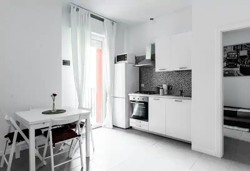 Angolo cottura moderno e luminoso con mobili bianchi, forno integrato e frigorifero, più un piccolo tavolo da pranzo accanto a una porta luminosa — perfetto per un appartamento cittadino compatto.