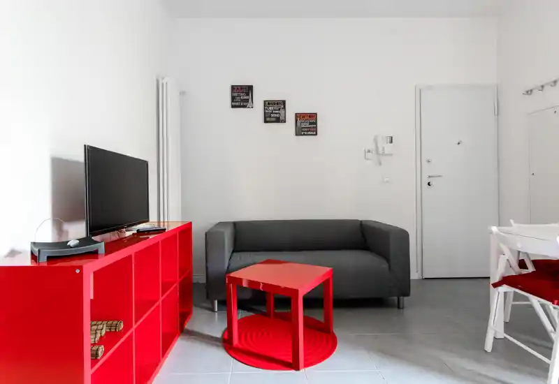 Soggiorno moderno e luminoso con un divano grigio compatto, scaffalatura rossa e tavolino coordinato che creano un look minimalista e deciso.
