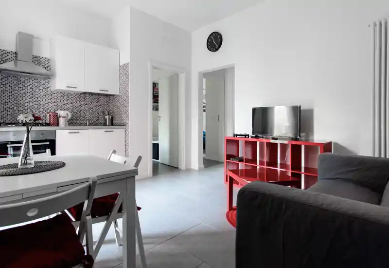 Zona giorno luminosa open-space con angolo cottura moderno, tavolo da pranzo e divano confortevole di fronte a una piccola TV su scaffalatura rossa — ideale per un appartamento cittadino.