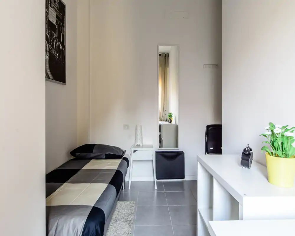 Dormitorio individual moderno y compacto con cama bien hecha, mesita y espejo de cuerpo entero; tonos neutros y mobiliario sencillo que crean un ambiente tranquilo para estancias cortas.