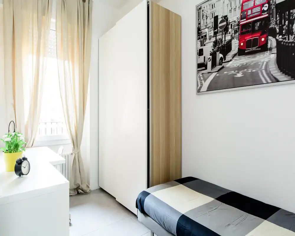 Dormitorio individual moderno y luminoso con cama bien hecha, escritorio blanco junto a la ventana y armario estrecho. Las cortinas suaves y el cuadro urbano aportan una sensación acogedora.