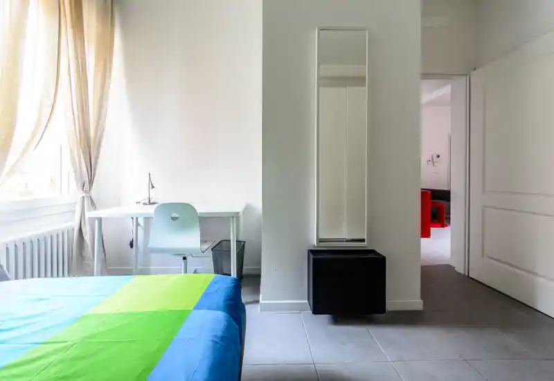 Dormitorio moderno y luminoso con colcha colorida, escritorio junto a la ventana y un espejo alto junto a la entrada — estilo limpio y mucha luz natural.