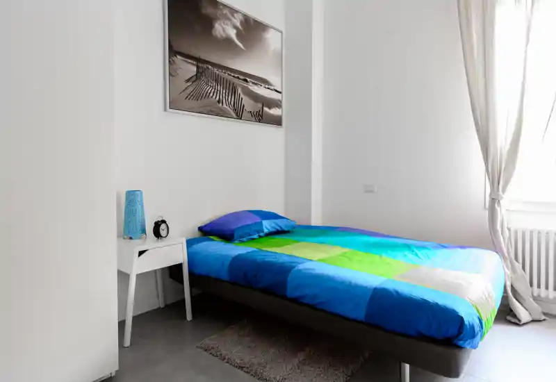 Dormitorio individual moderno y luminoso con ropa de cama colorida, mesita y luz natural de una gran ventana; estilo sencillo y limpio ideal para un anuncio.