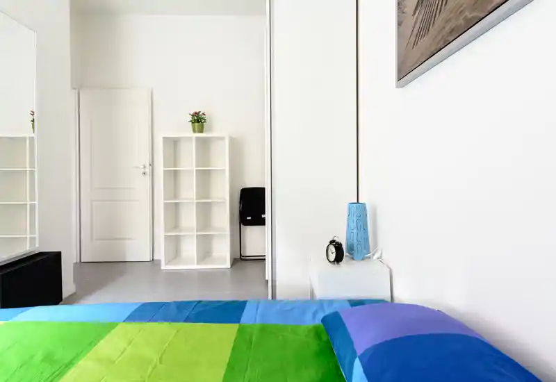 Dormitorio luminoso y minimalista con colcha colorida, mesita de noche y armario; líneas limpias y luz natural para una sensación moderna.