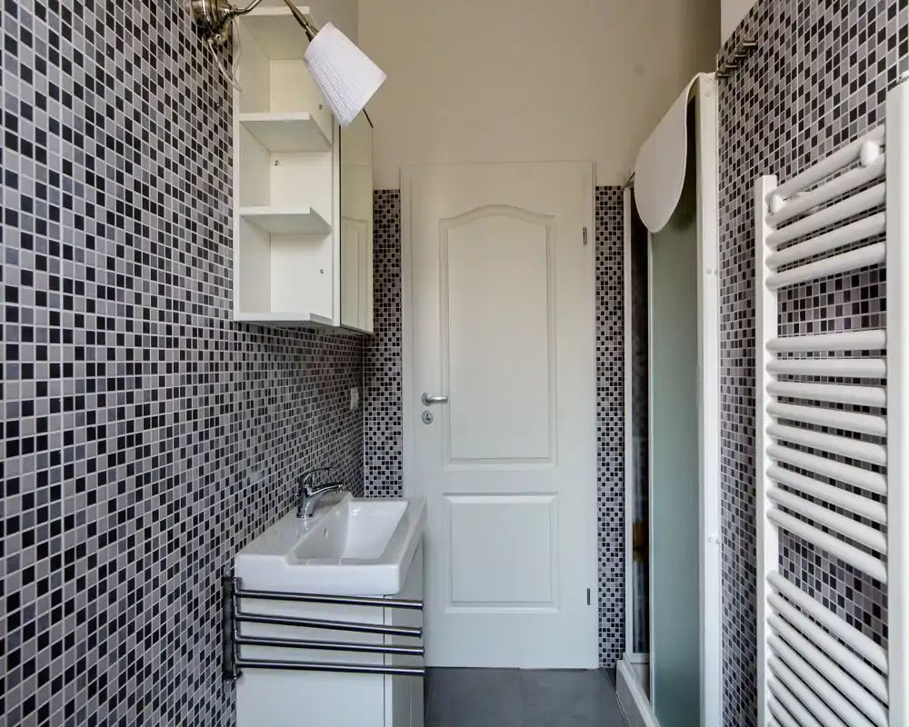 Baño moderno con azulejos en mosaico, lavabo, armario espejo, toallero radiador y cabina de ducha; acabados blancos y grises impecables.