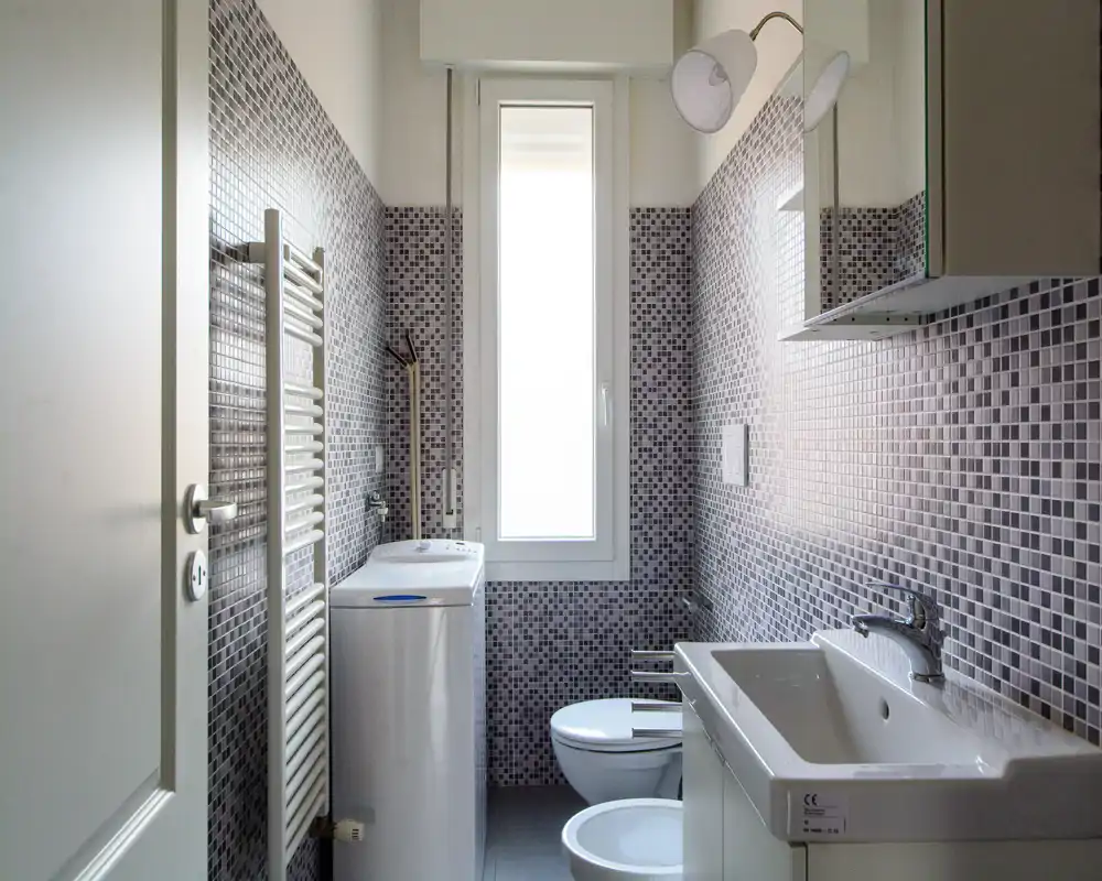 Baño moderno y limpio con paredes de mosaico, lavabo, bidé, inodoro y una lavadora junto a una ventana alta que aporta luz natural.