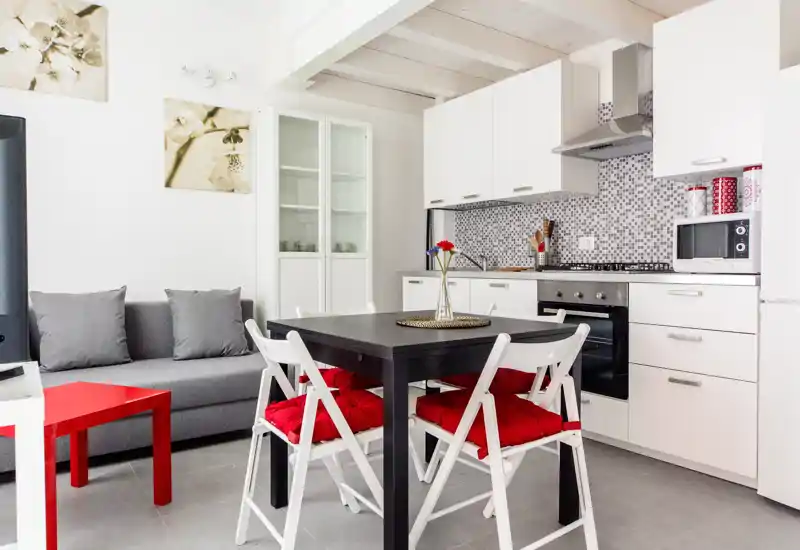 Zona pranzo luminosa e moderna adiacente a una cucina compatta, con tavolo nero, sedie bianche con cuscini rossi, divano grigio e mobili contemporanei—perfetta per annunci di monolocali o bilocali.