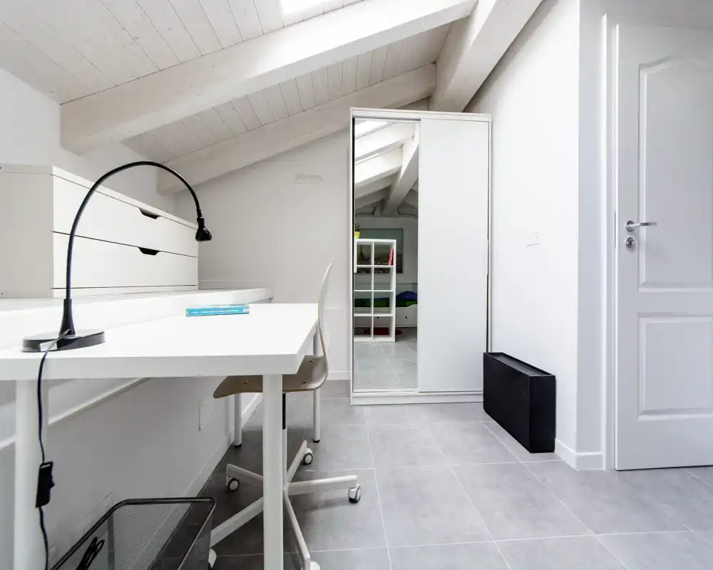 Spazio di lavoro in mansarda luminoso con scrivania bianca, lampada da tavolo e sedia — stile minimal e ordinato sotto il soffitto inclinato.