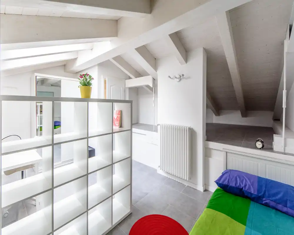 Camera da letto in mansarda luminosa con travi inclinate in legno, copriletto colorato e divisorio a scaffali bianco che definisce lo spazio; stile moderno, pulito e accogliente.