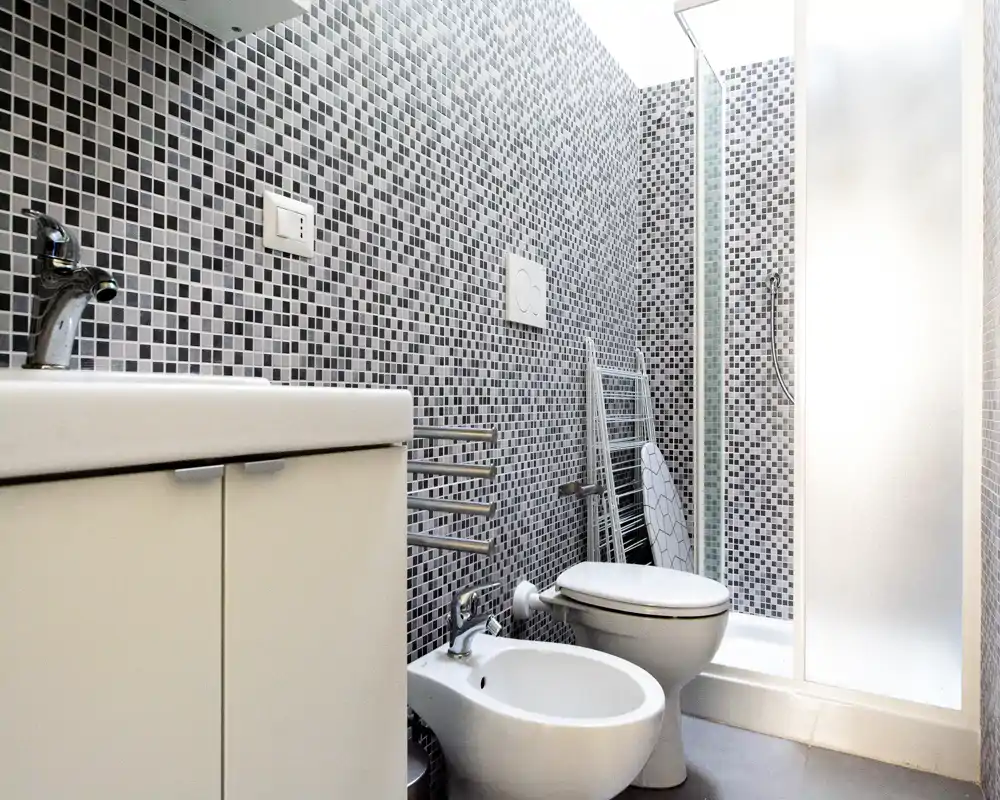 Bagno moderno compatto con pareti in mosaico, bidet, WC e cabina doccia. Design luminoso e pulito, ideale per appartamenti piccoli.