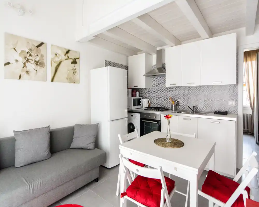 Cucina a vista luminosa e moderna con mobili bianchi, paraschizzi piastrellato, frigorifero, piano cottura e un piccolo tavolo da pranzo con sedie con cuscini rossi; un angolo living compatto con divano grigio completa l'ambiente.