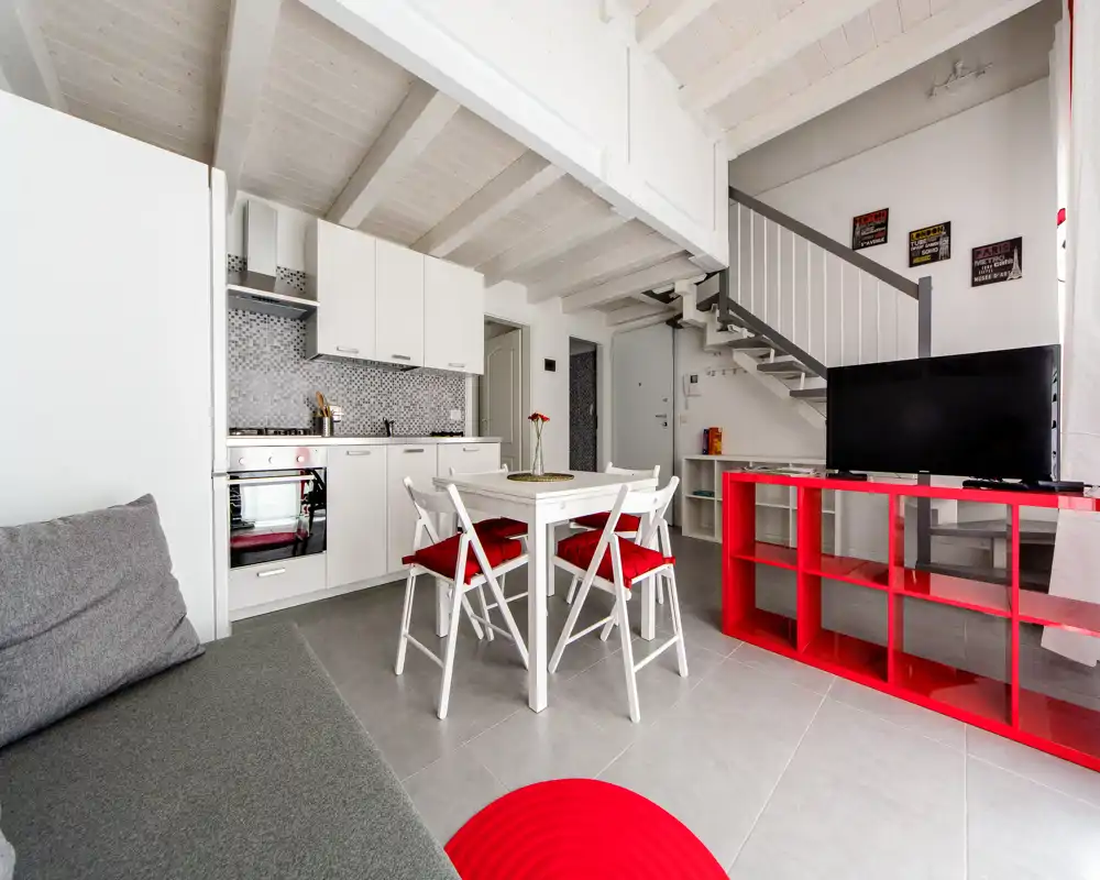 Soggiorno moderno e luminoso open‑space con angolo cottura compatto, tavolo da pranzo e zona TV; travi bianche e dettagli rossi per un tocco contemporaneo.