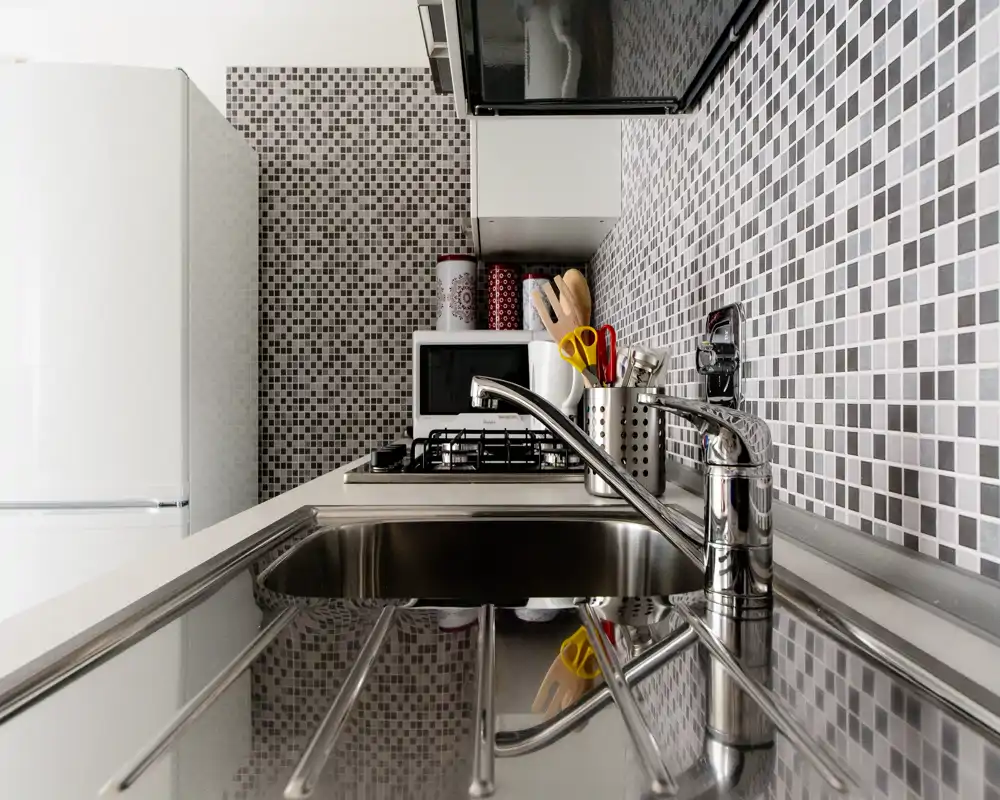 Cucina compatta e moderna con lavello in acciaio, schienale a mosaico, piano cottura a gas e piccoli elettrodomestici. Layout pulito ideale per affitti brevi.