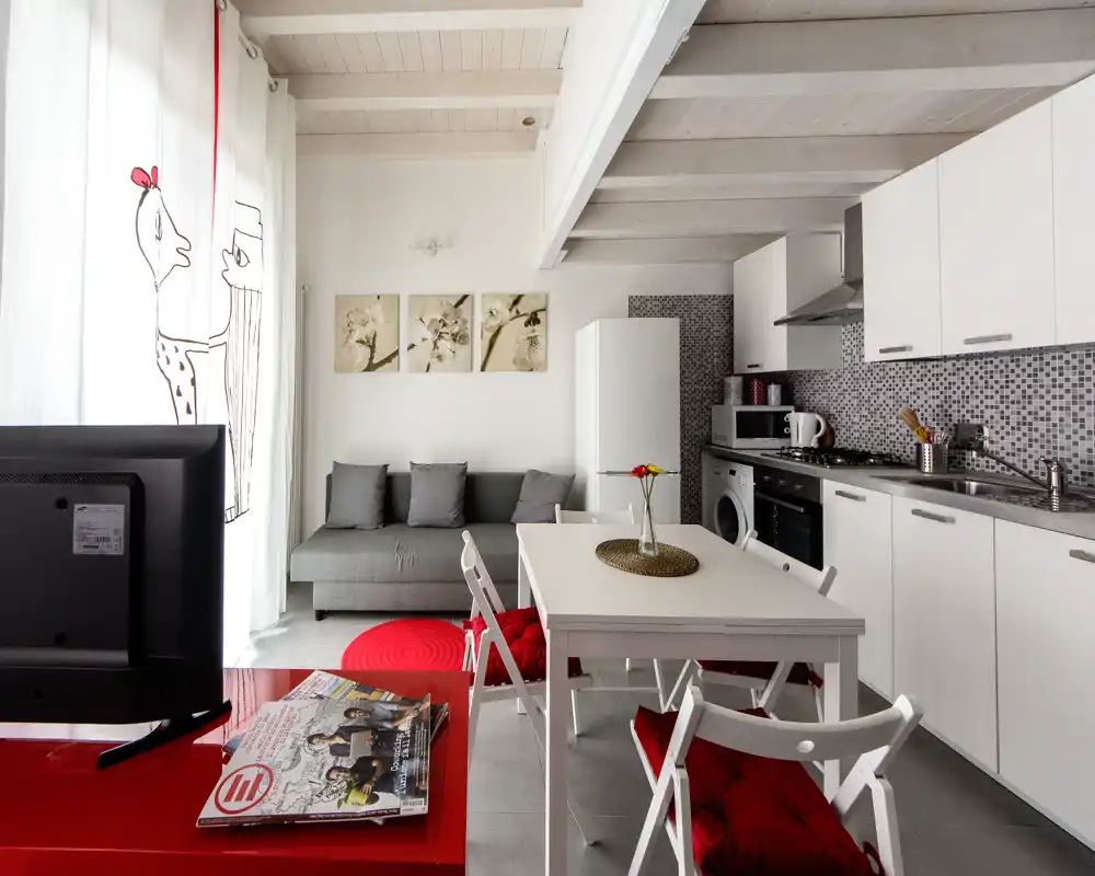 Zona giorno moderna e luminosa in open-space con cucina compatta, tavolo da pranzo e divano grigio — tocchi rossi e luce naturale rendono l'ambiente accogliente.