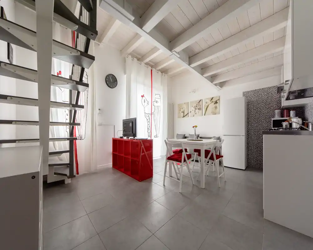 Zona giorno moderna e luminosa open‑space con tavolo da pranzo, mobile TV e angolo cottura visibile; arredo bianco contemporaneo con tocchi rossi e ampie finestre che garantiscono luce naturale.