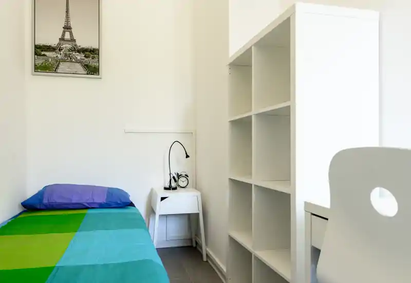 Dormitorio pequeño y luminoso con cama individual, mesita de noche con lámpara y despertador y estantería abierta — estilo minimalista y limpio, ideal para una persona.