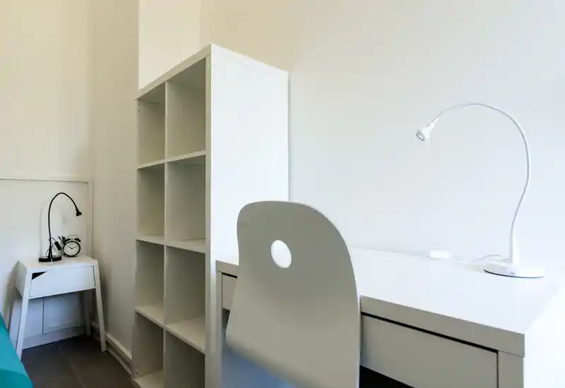 Rincón de dormitorio minimalista con escritorio blanco, estantería modular y mesita; una lámpara de escritorio y un despertador añaden funcionalidad a este espacio luminoso y neutro.