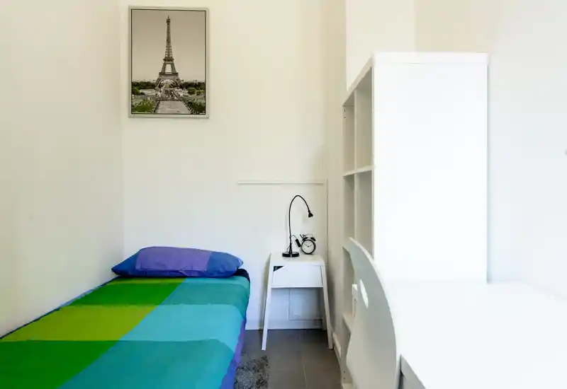 Habitación individual compacta con cama colorida, mesita y lámpara, estantería blanca y escritorio — luminosa, ordenada y minimalista.