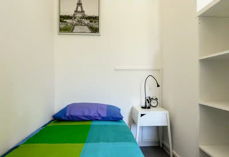 Habitación individual pequeña y luminosa con colcha colorida, mesita, lámpara y estanterías — ideal para un alquiler compacto.