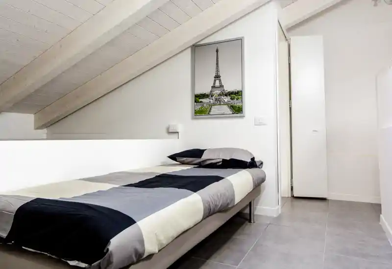 Camera da letto compatta e minimal con letto singolo sotto un soffitto spiovente, biancheria neutra e una stampa della Torre Eiffel incorniciata; luminosa, ben presentata e adatta a un monolocale in città.