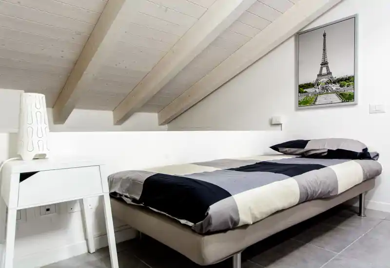 Camera mansardata compatta con letto singolo, biancheria dai toni neutri e travi in legno chiaro; luminosa, moderna e ordinata — ideale come affitto breve o stanza per gli ospiti.