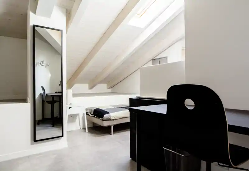 Camera da letto in mansarda luminosa con letto basso sotto travi bianche inclinate e una finestra a tetto; presente scrivania e specchio, arredamento minimal e moderno.