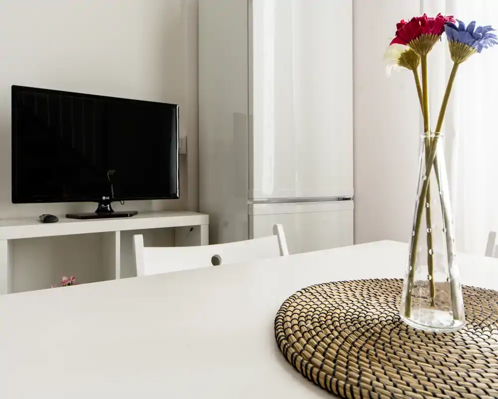 Zona pranzo luminosa e minimal con tavolo bianco, centro tavola in paglia e vaso di vetro con fiori; TV e frigorifero sullo sfondo, atmosfera da monolocale compatto.