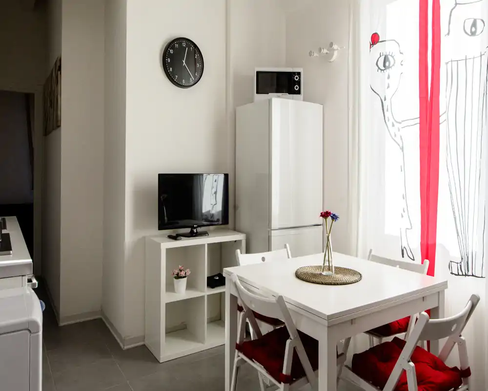 Zona pranzo compatta e luminosa con tavolo bianco e quattro sedie, cuscini rossi e un piccolo centrotavola; ampia finestra con tende decorate che dà luce naturale.