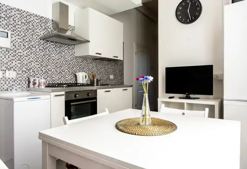 Cocina moderna y luminosa con muebles blancos, salpicadero de mosaico, horno empotrado y mesa con centro decorativo — área de cocina y comedor compacta y bien presentada.