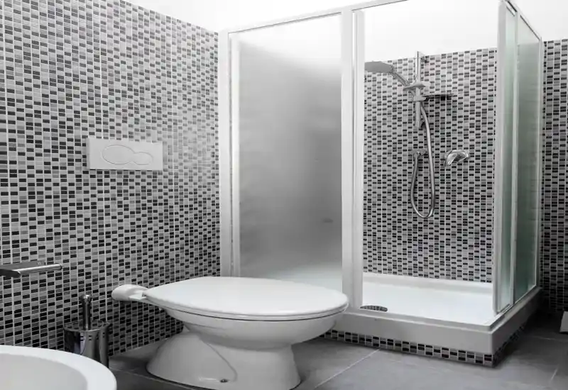 Bagno moderno e pulito con pareti in piastrelle a mosaico, box doccia con vetro satinato e WC bianco; ambiente luminoso e ben tenuto, ideale per annunci.