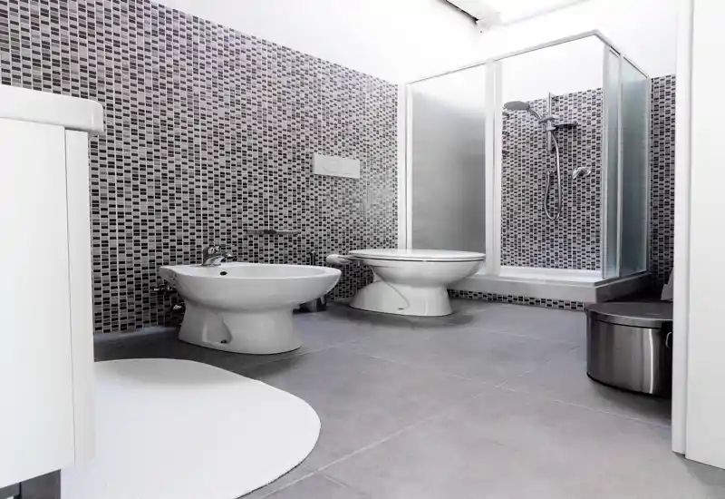 Bagno moderno con pareti piastrellate a mosaico, bidet e WC e box doccia in vetro; linee pulite e finiture neutre e luminose.