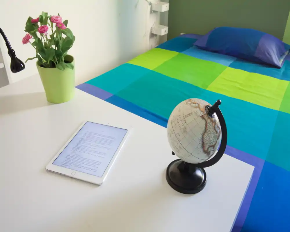 Bright, well-staged bedroom view with a colorful checkered bedspread, a pillow and a bedside desk showing a tablet, small globe and plant — ideal for modern rental listings.