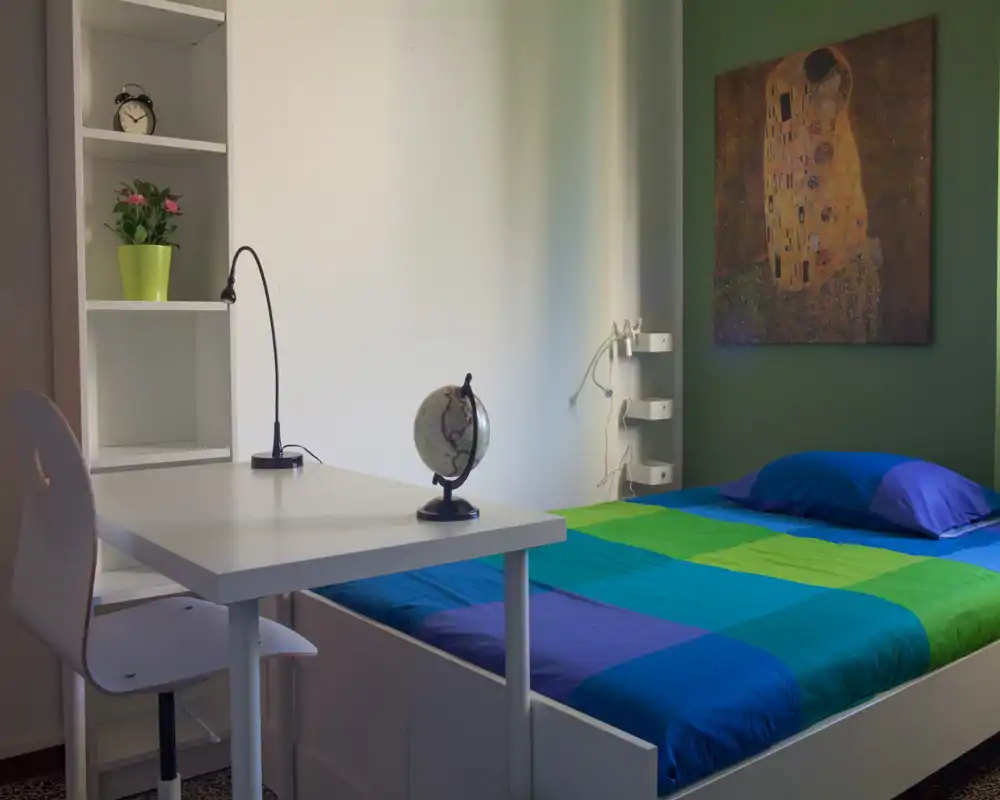 Bright, colorful bedroom with a single bed, neat desk and shelving. The room features a blue–green quilt, bedside wall art and a small workspace ideal for students or short-term rentals.