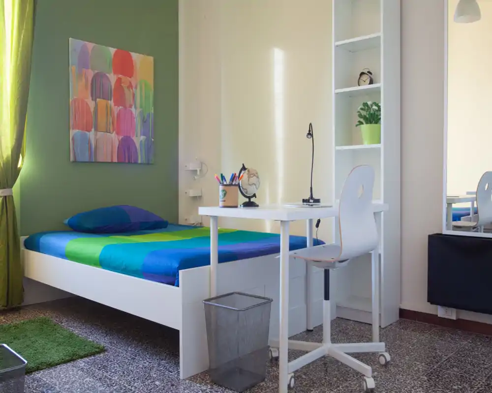 Bright, modern bedroom with a white bed and colorful blue-green bedding, a small white desk and shelving — ideal for a student or single occupant.