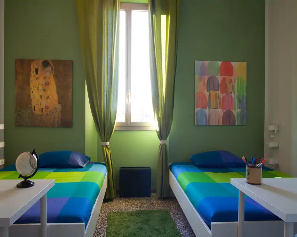 Bright twin bedroom with two single beds dressed in coordinated blue-green bedding, a central window with green curtains and colorful wall art — a tidy, welcoming space ideal for shared stays.