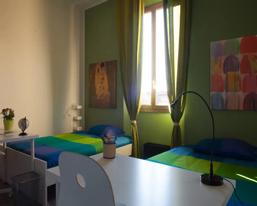 Bright, neatly staged twin-bedroom with colorful bedding, wall art and a desk in the foreground. Natural light from the central window and coordinating curtains create a warm, inviting atmosphere.