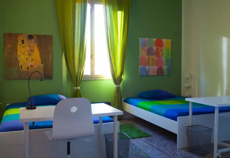 Camera doppia luminosa con due letti singoli, biancheria a righe colorate, scrivania e sedia davanti alla finestra, tende verdi e quadri decorativi. La luce naturale rende l'ambiente fresco e accogliente, perfetto per soggiorni brevi.